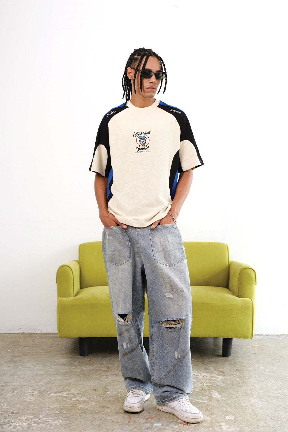 A model standing near a olive green couch with his hands in his pocket wearing Adventures Donald Duck Jersey Zoomed Out Front View from Bonkers Corner
